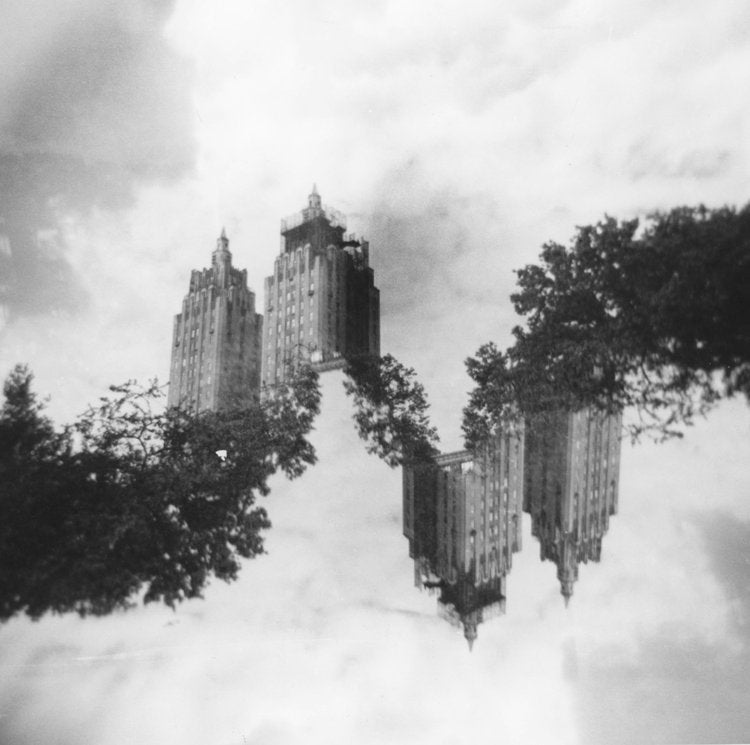 In a surreal composition, towering buildings seem to float amidst a cloudy sky, their mirrored...