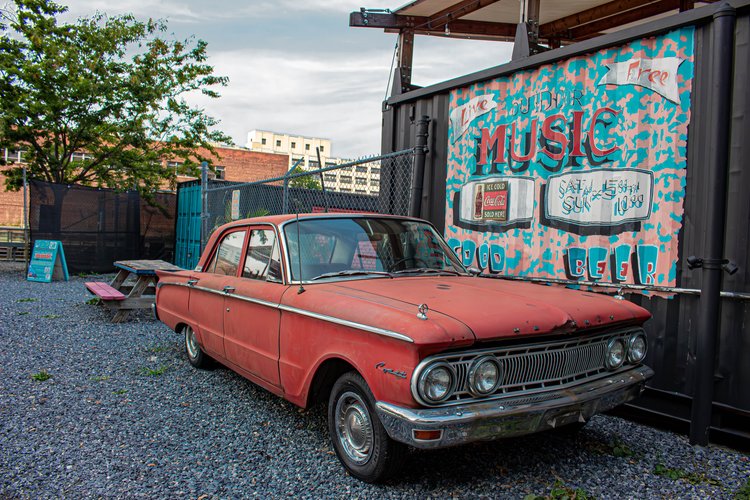 A vintage red car rests quietly against the backdrop of a vibrant mural, evoking nostalgia and a...